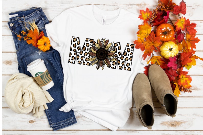May include: White t-shirt with the word "MOM" in black letters with a leopard print pattern. A sunflower with brown and yellow petals is in the center of the word.