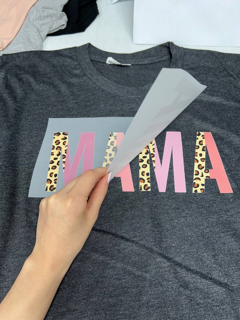 May include: Dark gray t-shirt with the word "MAMA" in pink and leopard print lettering. The image shows the application of the design to the shirt.