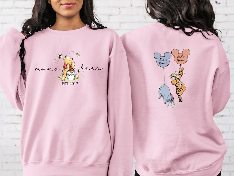Pu&ograve; includere: Una felpa rosa chiaro con Winnie the Pooh, il testo "mama bear" e "EST. 2012" sul davanti. Il retro della felpa mostra Eeyore e Tigro che tengono in mano dei palloncini con la scritta "Kid's Name".