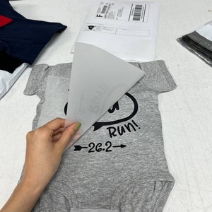 May include: A gray baby onesie with the text "Run!" and "26.2" in black. A hand is peeling away a transparent sheet from the onesie, revealing the design. Other packages are in the background.