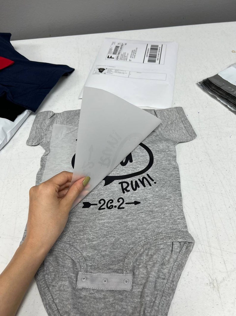 May include: A gray baby onesie with the text "Run!" and "26.2" in black. A hand is peeling away a transparent sheet from the onesie, revealing the design. Other packages are in the background.