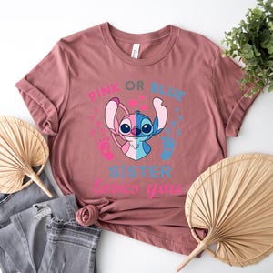 以下が含まれることがあります： ピンクかブルー？ハートと足跡がついたピンクとブルーのスティッチのキャラクター。テキストには「ピンクかブルー？妹はあなたを愛しています」と書かれています。シャツはダスティローズの色です。