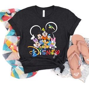 May include: Dark gray t-shirt featuring a colorful Disney character graphic. The graphic includes Mickey Mouse, Minnie Mouse, Donald Duck, and other characters. Pink flip-flops and a patterned scarf are also in the image.