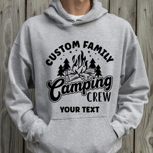 Pode incluir: Camisola de capuz cinzenta com um design gr&aacute;fico preto que diz "Custom Family Camping Crew" com uma fogueira e &aacute;rvores. O texto "Your Text" est&aacute; abaixo do gr&aacute;fico.