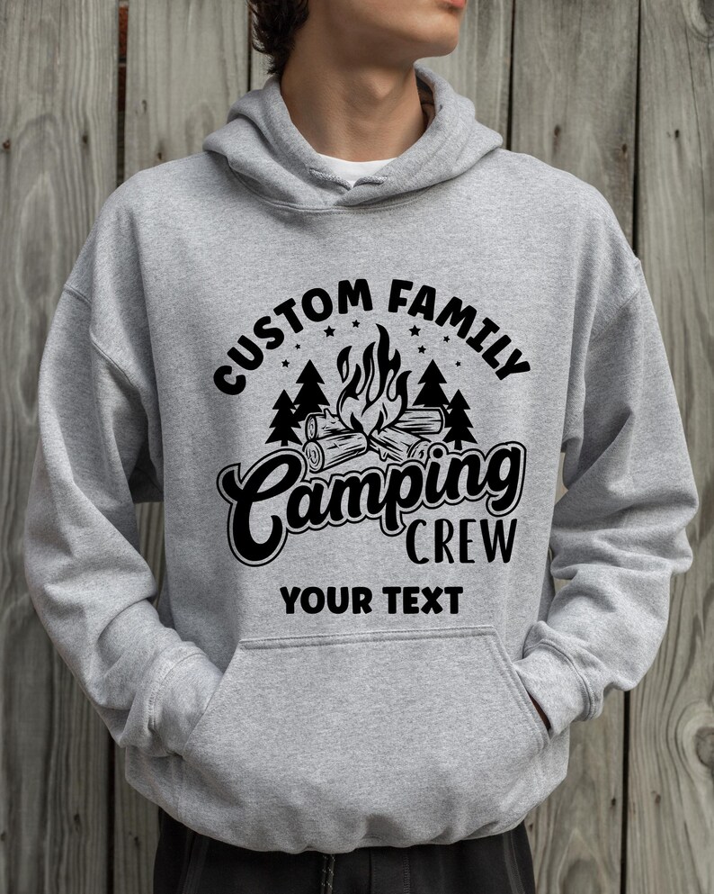 Pode incluir: Camisola de capuz cinzenta com um design gr&aacute;fico preto que diz "Custom Family Camping Crew" com uma fogueira e &aacute;rvores. O texto "Your Text" est&aacute; abaixo do gr&aacute;fico.
