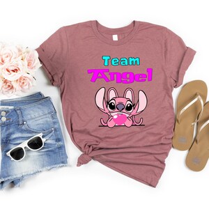 May include: A pink heather t-shirt with the text "Team Angel" in blue and pink lettering. A pink Stitch character with a heart is below the text.