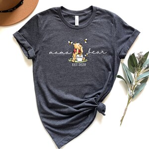 Pu&ograve; includere: T-shirt grigio scuro con una grafica di Winnie the Pooh che tiene un vasetto di miele con api. Il testo recita "mama bear" e "EST. 2020". La maglietta ha maniche arrotolate e orlo annodato.