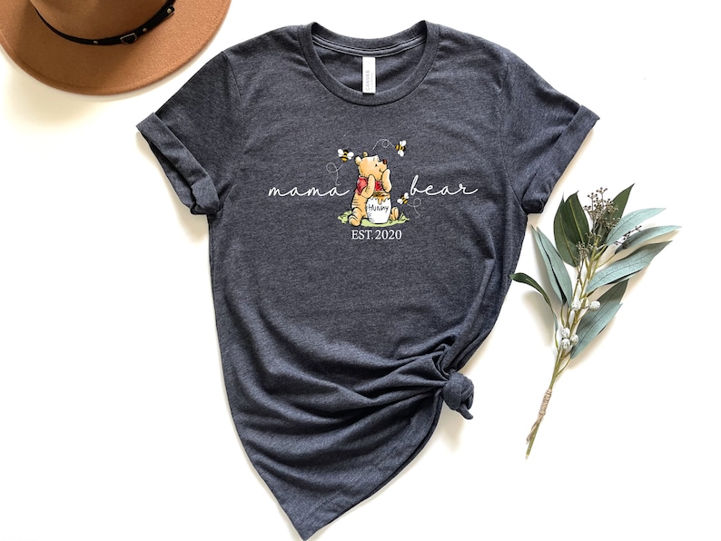 Pu&ograve; includere: T-shirt grigio scuro con una grafica di Winnie the Pooh che tiene un vasetto di miele con api. Il testo recita "mama bear" e "EST. 2020". La maglietta ha maniche arrotolate e orlo annodato.