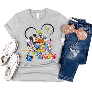 May include: A gray t-shirt featuring a colorful Disney character graphic, paired with blue jeans, white sneakers, and a rose gold sequined Minnie Mouse ear headband. The word "Disney" is printed in colorful letters.