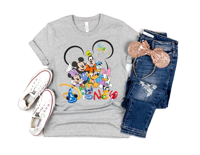 May include: A gray t-shirt featuring a colorful Disney character graphic, paired with blue jeans, white sneakers, and a rose gold sequined Minnie Mouse ear headband. The word "Disney" is printed in colorful letters.