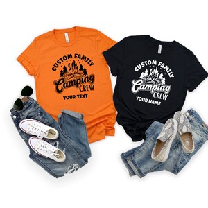 Pode incluir: Duas camisetas com tema de acampamento. A camiseta laranja tem texto preto que diz "Custom Family Camping Crew Your Text". A camiseta preta tem texto branco que diz "Custom Family Camping Crew Your Name". Ambas as camisetas apresentam um gr&aacute;fico de fogueira e &aacute;rvores.
