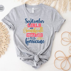 May include: Light grey t-shirt with the text "September Girls are sunshine mixed with a little hurricane" in blue, pink, and yellow.