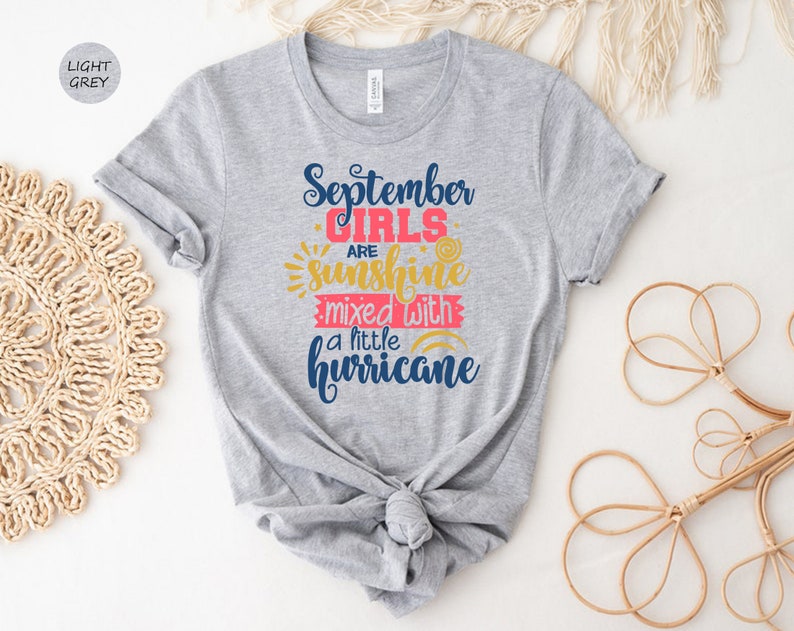 May include: Light grey t-shirt with the text "September Girls are sunshine mixed with a little hurricane" in blue, pink, and yellow.