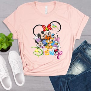 May include: A light pink t-shirt featuring a Disney design with Mickey Mouse, Minnie Mouse, Goofy, Donald Duck, Daisy Duck, and Pluto. The word "Disney" is written in colorful letters. White sneakers and blue jeans are also in the image.