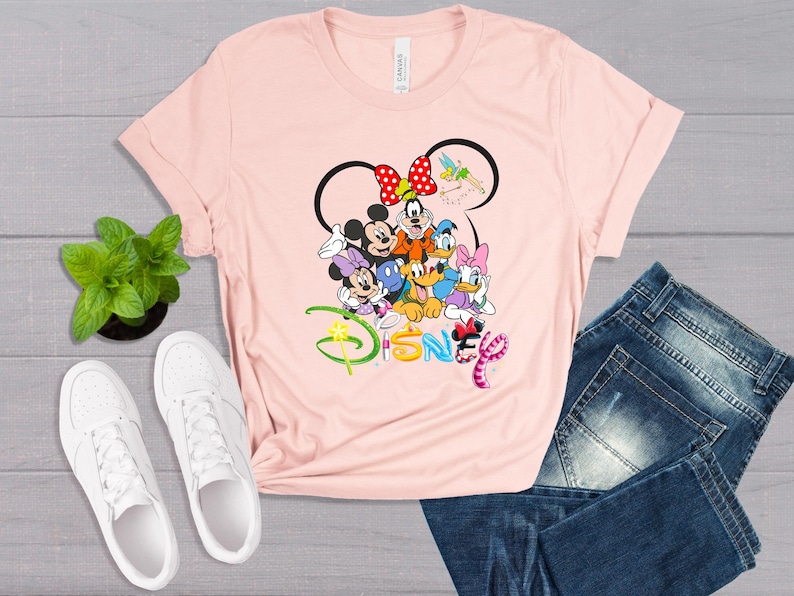 May include: A light pink t-shirt featuring a Disney design with Mickey Mouse, Minnie Mouse, Goofy, Donald Duck, Daisy Duck, and Pluto. The word "Disney" is written in colorful letters. White sneakers and blue jeans are also in the image.