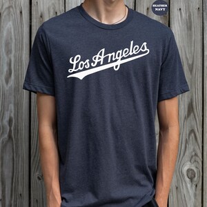 May include: A heather navy blue t-shirt with a white graphic of the text "Los Angeles" in a baseball-style font.
