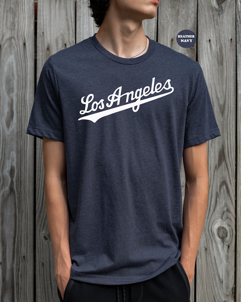 May include: A heather navy blue t-shirt with a white graphic of the text "Los Angeles" in a baseball-style font.