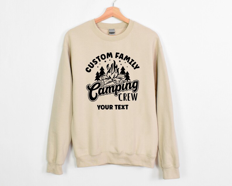 Pode incluir: Uma camisa de moletom bege com um design gr&aacute;fico preto que diz "Custom Family Camping Crew" com uma fogueira e &aacute;rvores. O texto "Your Text" est&aacute; abaixo do gr&aacute;fico.
