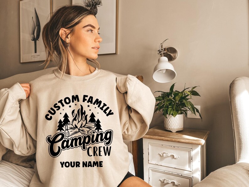 Pode incluir: Uma camisa de moletom bege com um design gr&aacute;fico preto que diz "Custom Family Camping Crew" e "Your Name".