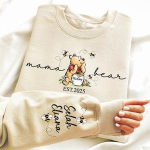 May include: A beige sweatshirt with the text "mama bear" and a cartoon Winnie the Pooh holding a honey pot. The text "EST. 2025" is below the image. The sleeve of the sweatshirt has the text "Selah Eliana" in black.