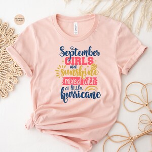 May include: A pink t-shirt with the text "September girls are sunshine mixed with a little hurricane" in blue and red lettering with a sun and rainbow graphic.