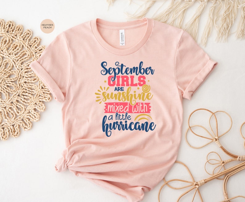 May include: A pink t-shirt with the text "September girls are sunshine mixed with a little hurricane" in blue and red lettering with a sun and rainbow graphic.