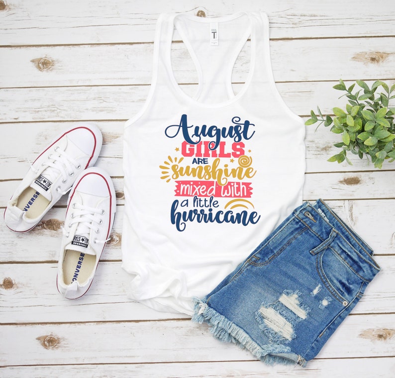 May include: White tank top with the text "August girls are sunshine mixed with a little hurricane" in pink, blue, and gold lettering. The tank top is laying on a white wooden surface with a pair of white sneakers and blue denim shorts.