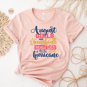 May include: A pink t-shirt with the text "August Girls are sunshine mixed with a little hurricane" in blue and pink lettering. The text is surrounded by yellow stars and a rainbow.