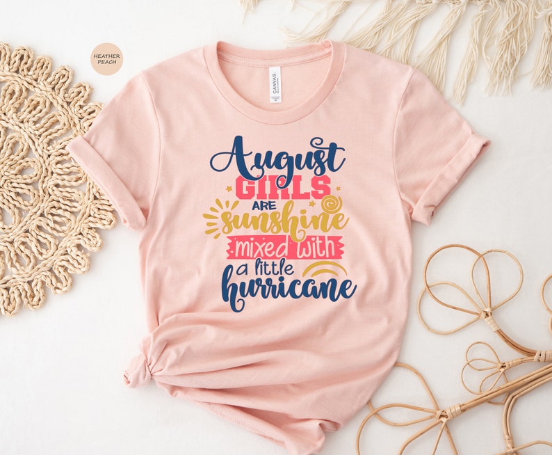 May include: A pink t-shirt with the text "August Girls are sunshine mixed with a little hurricane" in blue and pink lettering. The text is surrounded by yellow stars and a rainbow.