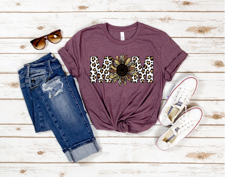 May include: A maroon t-shirt with the word "MOM" in large letters. The letters are decorated with a leopard print pattern and a sunflower graphic.