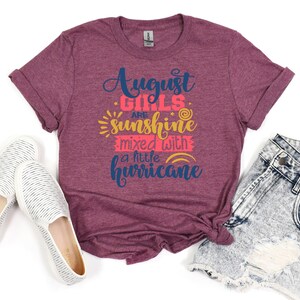 May include: A maroon t-shirt with the text "August Girls are sunshine mixed with a little hurricane" in navy blue, pink, and yellow. The shirt is folded and laid on a light blue denim shorts and two white canvas shoes.