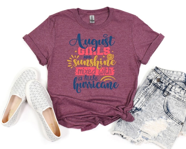 May include: A maroon t-shirt with the text "August Girls are sunshine mixed with a little hurricane" in navy blue, pink, and yellow. The shirt is folded and laid on a light blue denim shorts and two white canvas shoes.