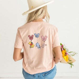 Pu&ograve; includere: T-shirt color pesca chiaro con tre personaggi di Winnie the Pooh sospesi a palloncini a forma di orecchie di Topolino. Ogni palloncino riporta la scritta "Kid's Name". I personaggi sono Pooh, Eeyore e Pimpi.