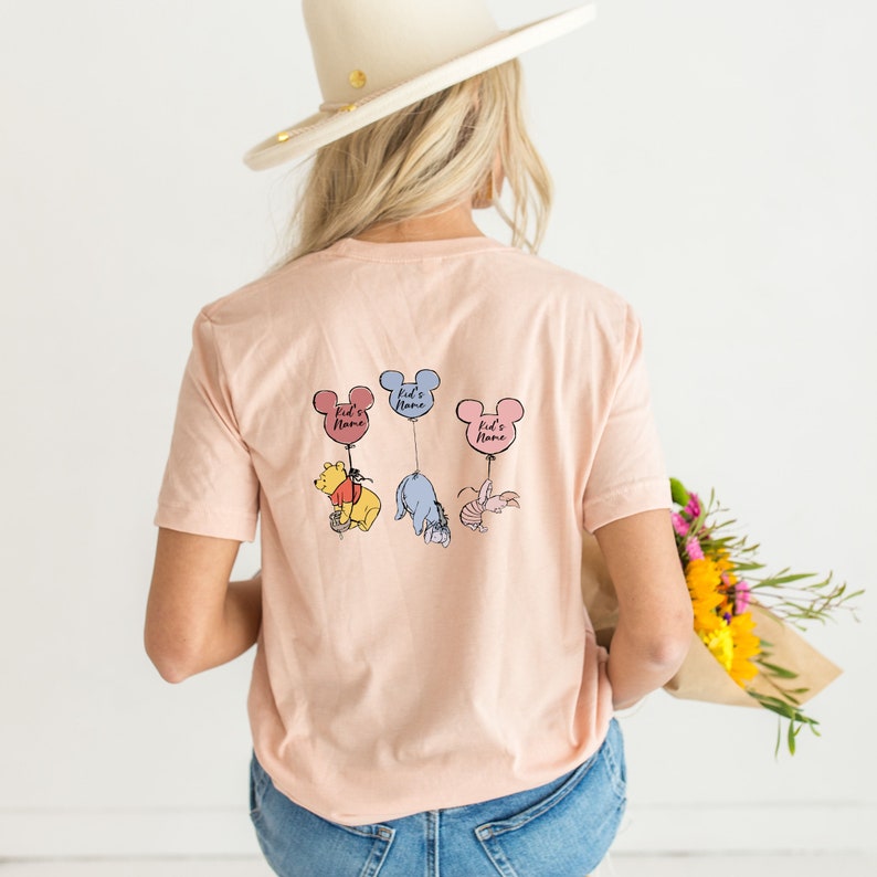 Pu&ograve; includere: T-shirt color pesca chiaro con tre personaggi di Winnie the Pooh sospesi a palloncini a forma di orecchie di Topolino. Ogni palloncino riporta la scritta "Kid's Name". I personaggi sono Pooh, Eeyore e Pimpi.
