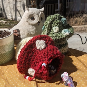 May include: A red, cream, and green crocheted mushroom dice bag with a drawstring closure. The bag is decorated with white crocheted flower accents. Several dice are visible inside and next to the bag. An owl figurine is in the background.