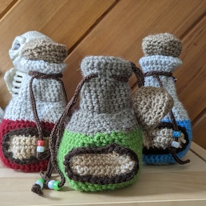 Könnte beinhalten: Drei gehäkelte Zaubertrankflaschen in Rot, Grün und Blau. Jede Flasche hat einen gehäkelten Stopfen und ein gehäkeltes Etikett in Braun.