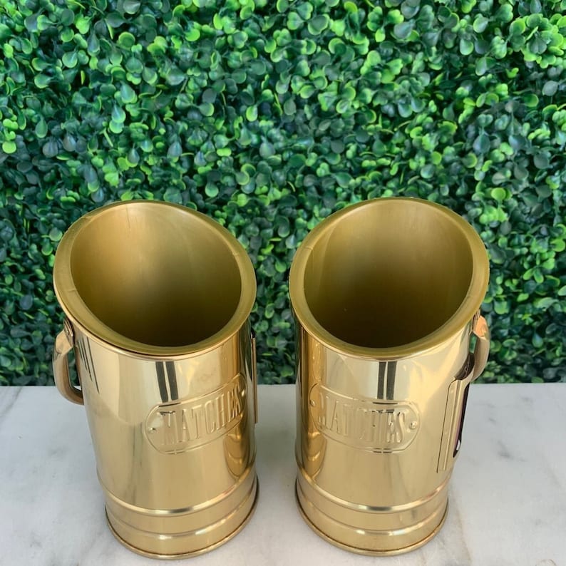 May include: Two gold metal containers with a slanted top and a handle on the side. The containers are labelled "Matches" and "Candles".