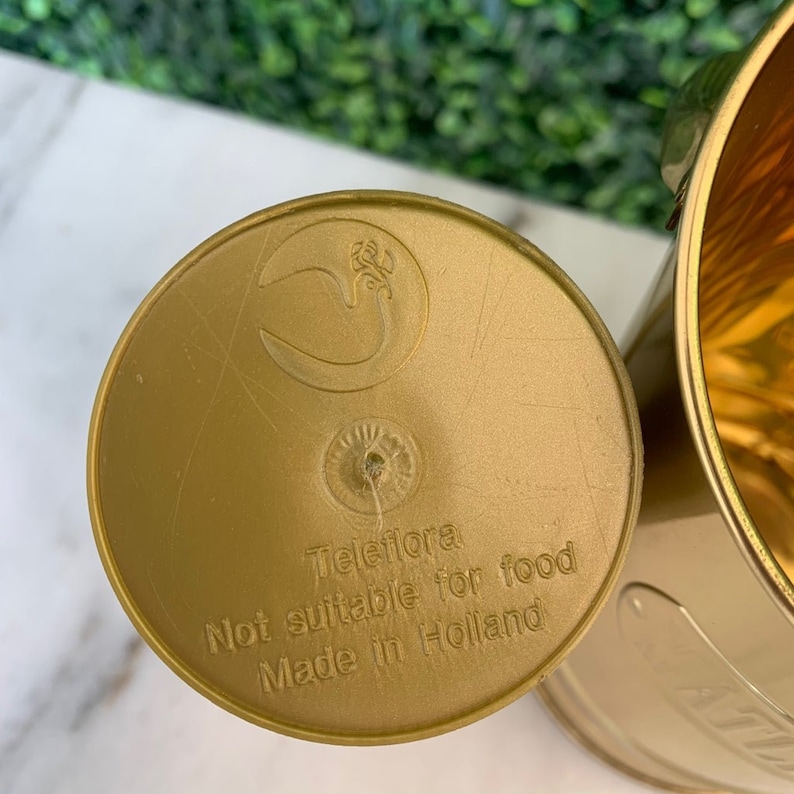 May include: Gold metal container with a lid. The lid has a logo and the text "Teleflora Not suitable for food Made in Holland".
