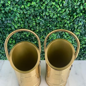 May include: Two gold metal bottle holders with the word "Matches" printed on the front. The holders are empty and have a shiny finish.