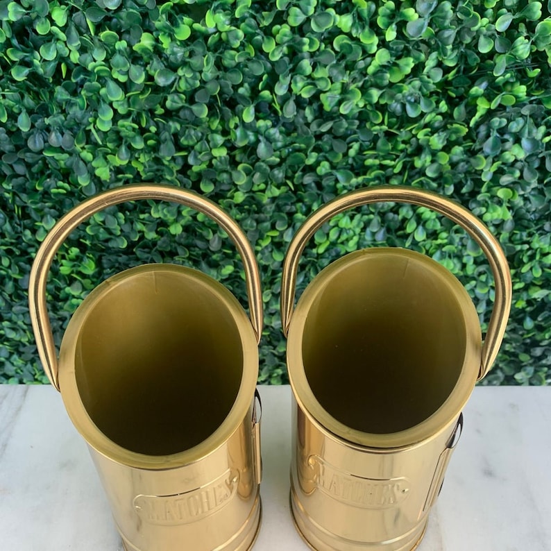 May include: Two gold metal bottle holders with the word "Matches" printed on the front. The holders are empty and have a shiny finish.