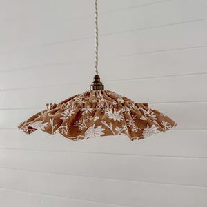 Puede incluir: Una pantalla de lámpara con estampado floral marrón y blanco con un borde con volantes cuelga de un cable blanco. La pantalla de la lámpara está suspendida de un accesorio de metal oscuro. El fondo es una pared blanca con paneles de madera horizontales.