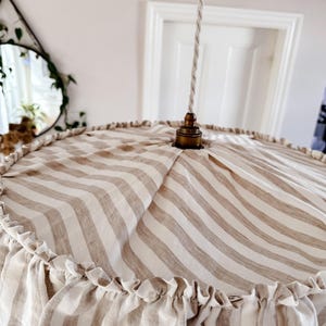 May include: A beige and white striped lampshade with a ruffled edge. The lampshade is made of fabric and has a simple, elegant design.