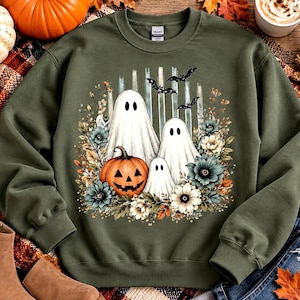 May include: An olive green crewneck sweatshirt featuring a graphic print of three white ghosts, a carved orange jack-o'-lantern, and various autumn-toned flowers. The design includes vertical white brushstrokes and three black flying bats.