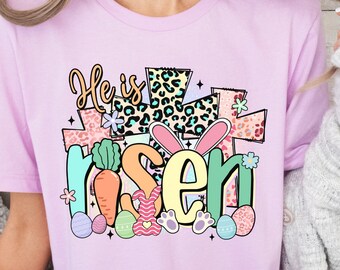 May include: A pink t-shirt with a colorful graphic design that says "He is risen" with a cross, Easter eggs, and a bunny rabbit.