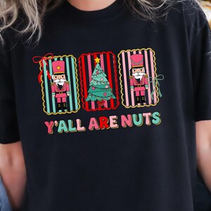 May include: Black t-shirt featuring a festive design with two nutcrackers and a Christmas tree. The design includes the text "Y'all Are Nuts" in colorful lettering, perfect for the holiday season.