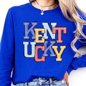 May include: A royal blue sweatshirt with the word "KENTUCKY" in a colorful, stacked design. The letters are outlined in black and filled with shades of yellow, orange, blue, and red. The sweatshirt is a solid color.