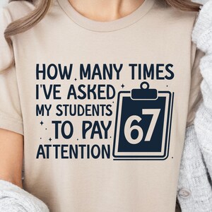 May include: A beige t-shirt with the text "HOW MANY TIMES I'VE ASKED MY STUDENTS TO PAY ATTENTION" in dark blue. A dark blue clipboard graphic displays the number "67".