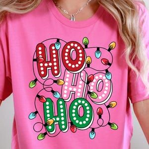 May include: Pink t-shirt with a festive "HO HO HO" design. The text is outlined in white and filled with red, pink, and green, and decorated with colorful Christmas lights. A fun and cheerful holiday tee.