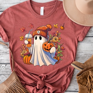 May include: A red t-shirt with a cartoon ghost wearing a pumpkin-colored hat and holding a pumpkin and a cup of coffee. The ghost is surrounded by fall leaves and flowers.