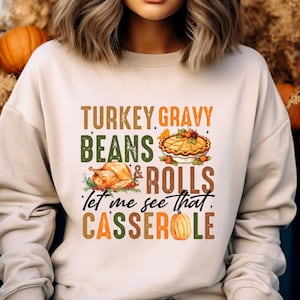 May include: Cream-colored sweatshirt with a Thanksgiving-themed graphic. The design includes text that reads "TURKEY GRAVY BEANS & ROLLS Let me see that CASSEROLE" along with illustrations of a turkey, pie, and pumpkin.